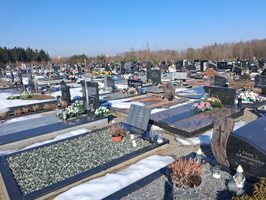 Wide cemetery scene with rows of gravestones, flowers, and candles, snow on the ground under a clear blue sky.